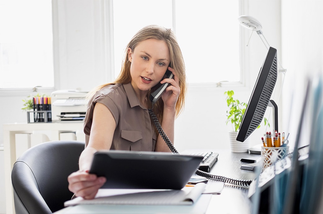 En kvinna som arbetar vid sitt skrivbord och tittar på en surfplatta medan hon talar i telefon.